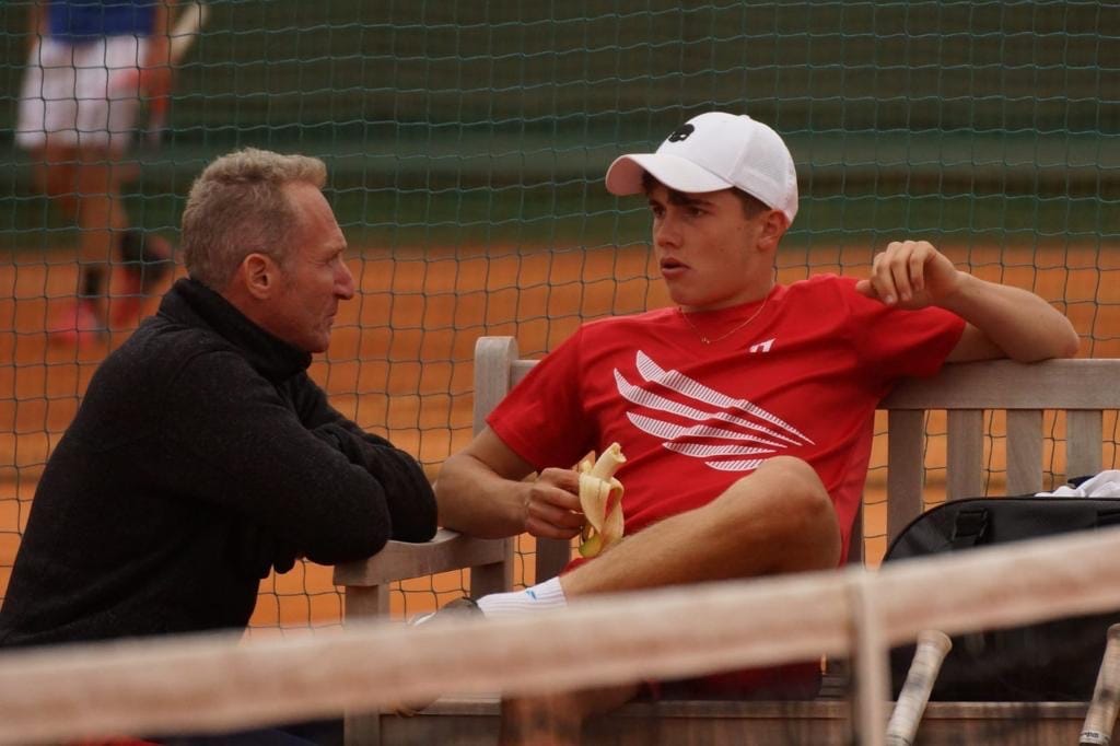 Benoit Foucher talking with a player on a bench beside a clay court